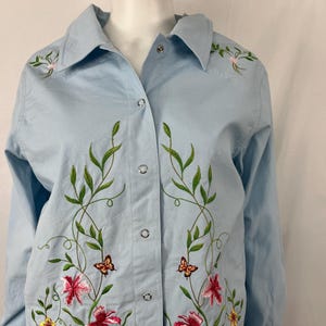 May include: Light blue button-up shirt with floral embroidery. The shirt features a collar and long sleeves. The embroidery includes pink, red, and yellow flowers, green leaves, and butterflies.
