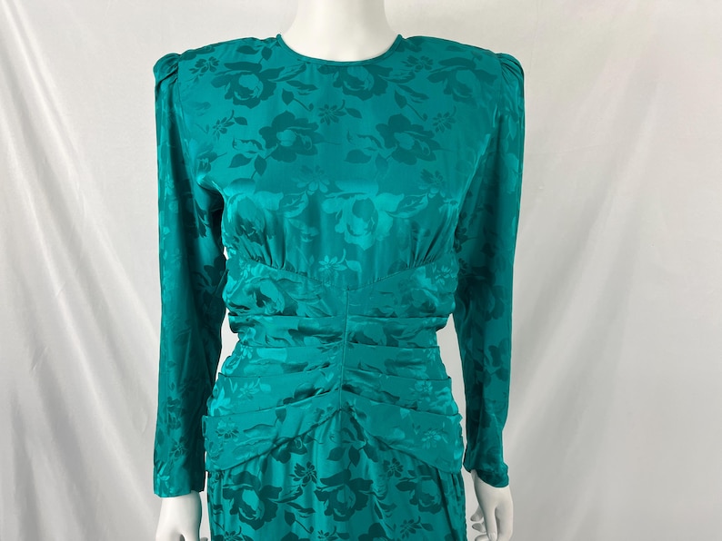 May include: Teal-colored vintage dress with a floral pattern. The dress features long sleeves, shoulder pads, and a ruched waist and skirt. The fabric has a subtle sheen, adding to its elegant appearance. A classic, formal dress.