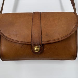 May include: A vintage brown leather crossbody bag with a flap closure and a gold-tone clasp. The bag has a rectangular shape with rounded edges and a long, thin strap. The leather shows signs of wear, adding to its classic appeal.