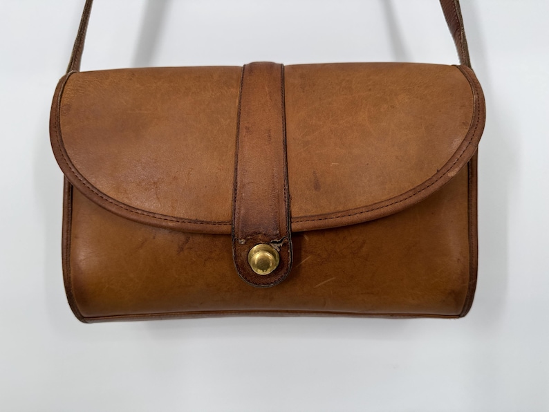 May include: A vintage brown leather crossbody bag with a flap closure and a gold-tone clasp. The bag has a rectangular shape with rounded edges and a long, thin strap. The leather shows signs of wear, adding to its classic appeal.