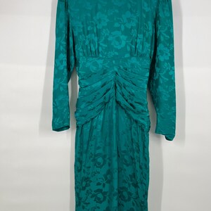 May include: A teal green vintage dress with long sleeves and a floral pattern. The dress features a ruched bodice and a knee-length skirt. The dress is hanging on a white hanger against a white background.