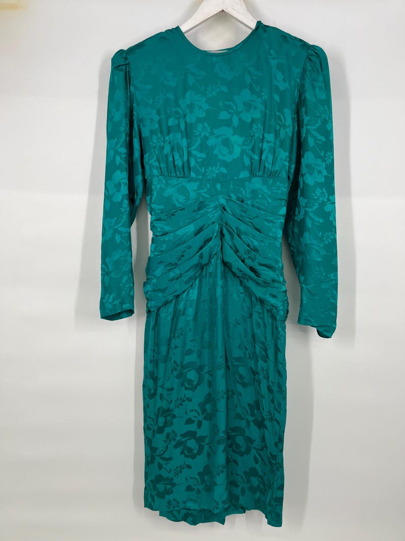 May include: A teal green vintage dress with long sleeves and a floral pattern. The dress features a ruched bodice and a knee-length skirt. The dress is hanging on a white hanger against a white background.