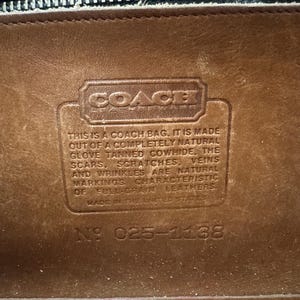 May include: Close-up of a brown leather Coach bag interior. The Coach logo is visible, along with text stating the bag is made of natural glove-tanned cowhide. The text also mentions natural markings and full-grain leather. The serial number is 025-1138.