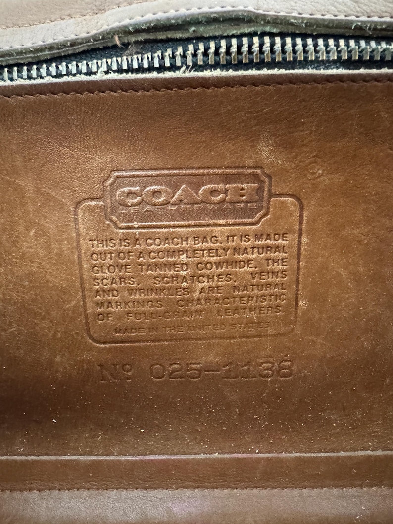 May include: Close-up of a brown leather Coach bag interior. The Coach logo is visible, along with text stating the bag is made of natural glove-tanned cowhide. The text also mentions natural markings and full-grain leather. The serial number is 025-1138.