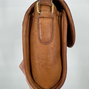 May include: A brown leather crossbody bag with a flap closure and adjustable strap. The bag features a brass-colored buckle and a worn, textured appearance. The bag is held by a person's hand.