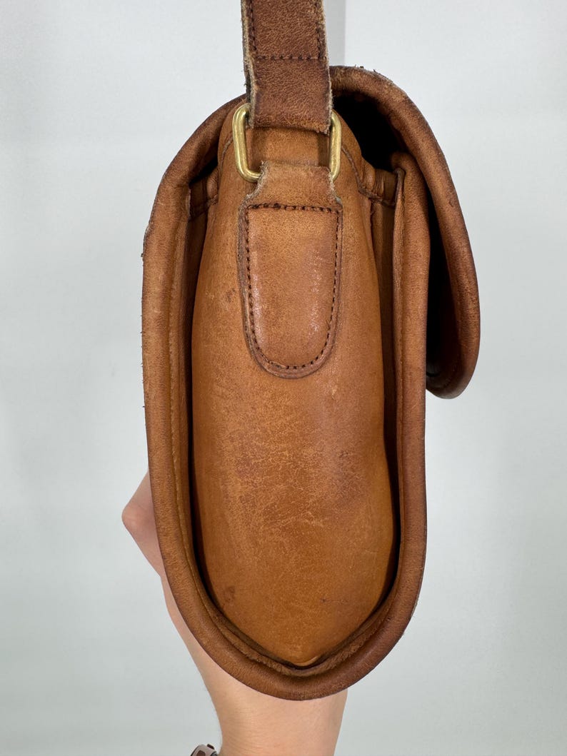 May include: A brown leather crossbody bag with a flap closure and adjustable strap. The bag features a brass-colored buckle and a worn, textured appearance. The bag is held by a person's hand.