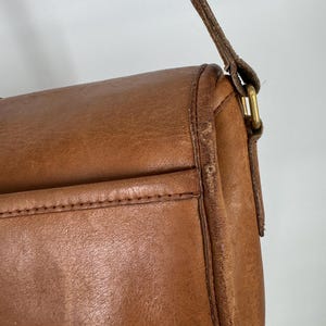 May include: Close-up of a brown leather crossbody bag. The bag features a flap closure, a stitched pocket, and a long, thin strap. The leather has a worn appearance, with visible grain and slight discoloration.