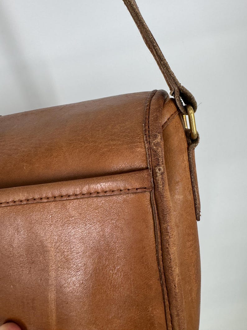May include: Close-up of a brown leather crossbody bag. The bag features a flap closure, a stitched pocket, and a long, thin strap. The leather has a worn appearance, with visible grain and slight discoloration.