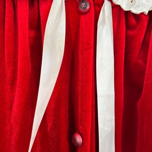 Op de afbeelding: Close-up van een rode fluwelen badjas met witte kanten afwerking en witte satijnen lintaccenten. De badjas heeft decoratieve knopen en een zachte, luxueuze textuur, wat wijst op een vintage of elegant ontwerp.