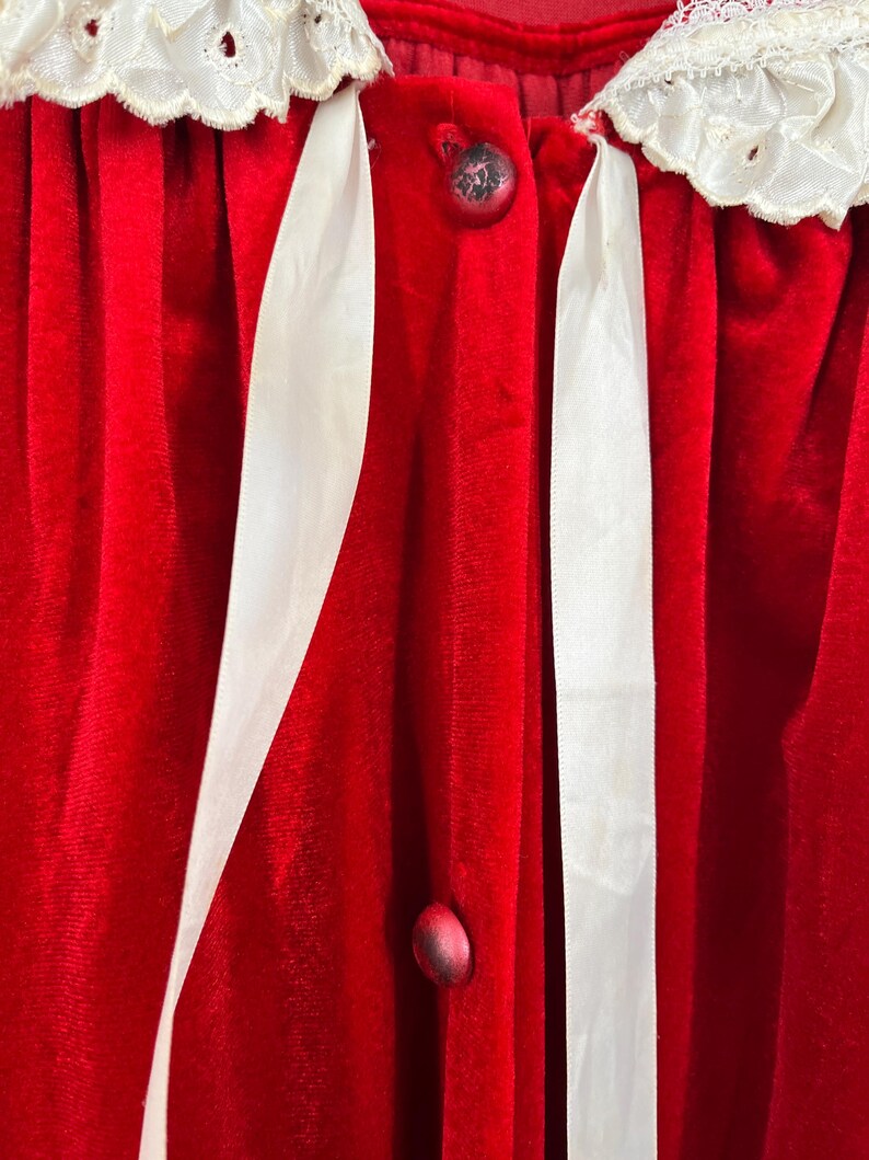 Op de afbeelding: Close-up van een rode fluwelen badjas met witte kanten afwerking en witte satijnen lintaccenten. De badjas heeft decoratieve knopen en een zachte, luxueuze textuur, wat wijst op een vintage of elegant ontwerp.