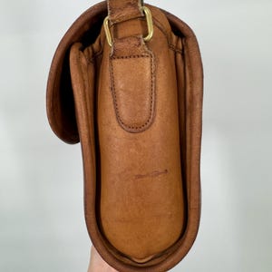 May include: A brown leather crossbody bag with a flap closure and adjustable strap. The bag features brass-colored hardware and visible stitching details. The leather has a worn appearance, suggesting vintage style.
