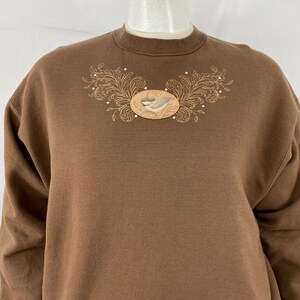May include: A brown sweatshirt with a crew neck. The front features an embroidered design of a bird and floral elements. The embroidery is in shades of tan, brown, and white. The sweatshirt is made of a soft material.