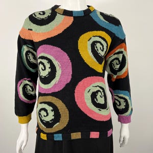 May include: Black sweater with colorful spiral designs in pink, yellow, green, orange, and blue. The long-sleeved sweater has a crew neck and ribbed cuffs. The bottom hem has a striped design.