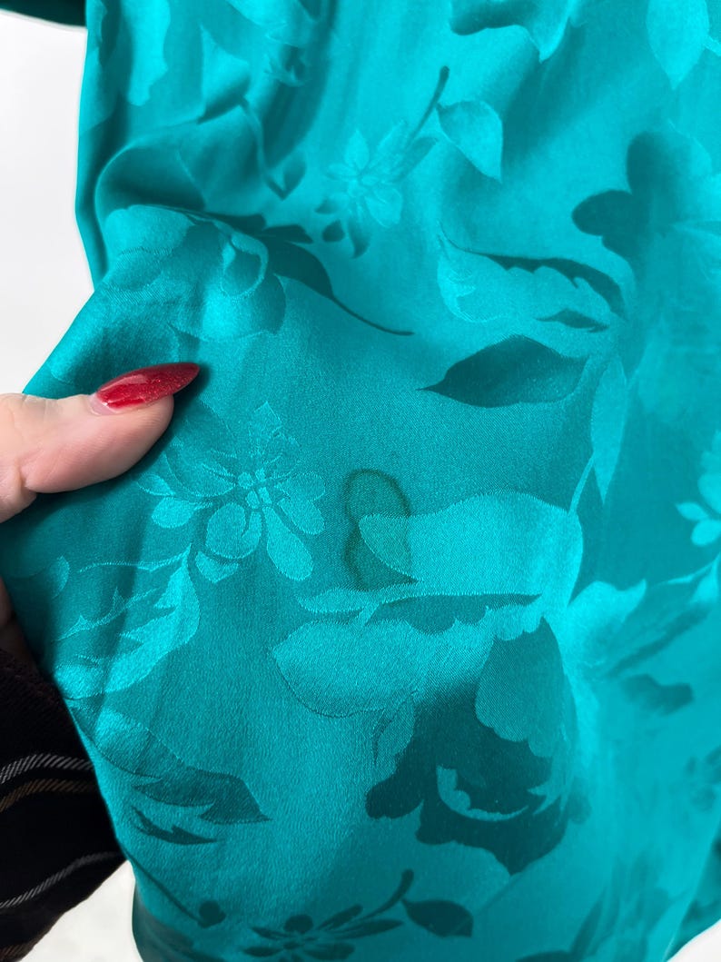 May include: Teal-colored fabric with a floral pattern. The fabric has a sheen and a soft texture. The floral design is a darker shade of teal, creating a subtle contrast. The fabric is gathered, showing its drape.