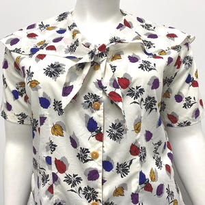 May include: A short-sleeved white blouse featuring a colorful leaf and floral print in shades of red, blue, purple, and yellow. The garment includes a wide collar with a tie-front detail and a vertical row of four round, light-colored buttons.