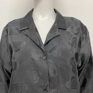 May include: A dark gray button-down shirt with a heart pattern. The long-sleeved shirt has a collared neckline and is made of a silky material. The heart pattern is subtly embossed throughout the fabric.