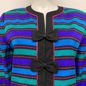 May include: A colorful striped jacket with a round collar and long sleeves. The jacket features horizontal stripes in blue, teal, purple, and pink, with black accents and two black bow details.
