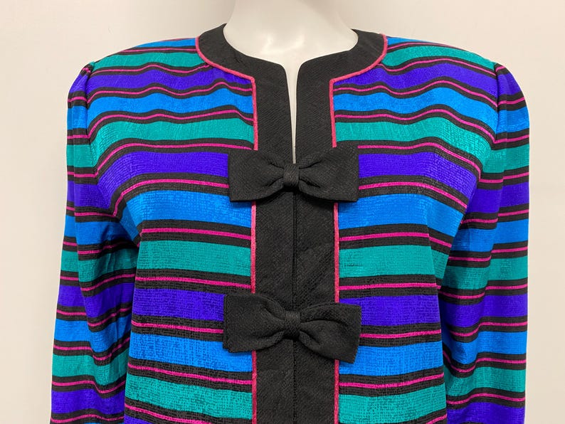 May include: A colorful striped jacket with a round collar and long sleeves. The jacket features horizontal stripes in blue, teal, purple, and pink, with black accents and two black bow details.