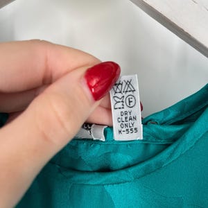 May include: Close-up of a teal-colored garment with a floral pattern. A white tag with laundry symbols and the text "DRY CLEAN ONLY K-555" is visible. The fabric appears to be satin or a similar material.