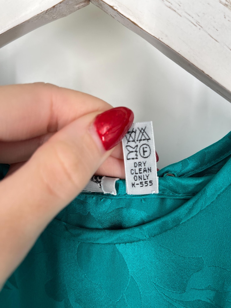 May include: Close-up of a teal-colored garment with a floral pattern. A white tag with laundry symbols and the text "DRY CLEAN ONLY K-555" is visible. The fabric appears to be satin or a similar material.