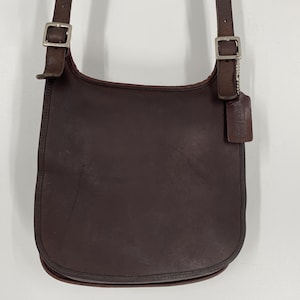 May include: A dark brown leather crossbody bag with a rounded bottom, featuring an adjustable shoulder strap with silver-toned metal buckles and a rectangular leather hang tag attached to the right side of the strap hardware.