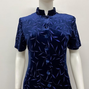 May include: A dark blue velvet dress featuring a mandarin collar, short sleeves, and a vertical row of matching fabric-covered buttons. The fabric is adorned with a repeating pattern of shimmering blue leaves and thin stems throughout the garment.