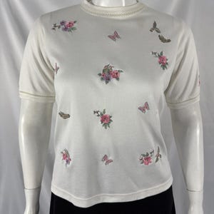 May include: Cream-colored short-sleeved shirt with embroidered floral and butterfly designs. The crew neck and sleeve hems have a delicate trim. The shirt is displayed on a mannequin, paired with a black skirt.