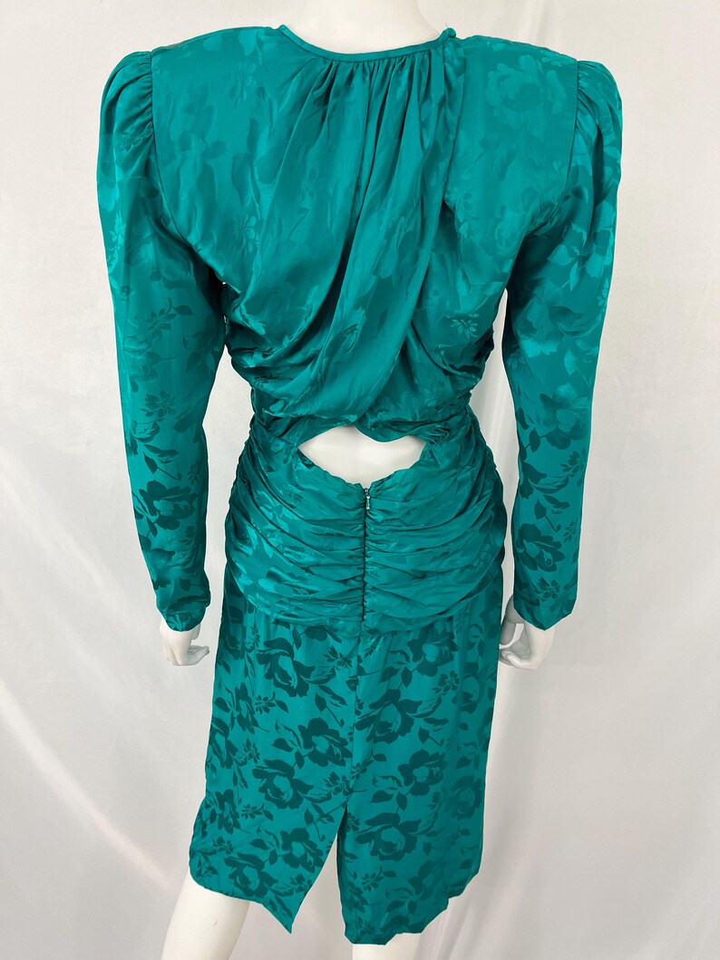 May include: Teal-colored vintage dress with a floral pattern. The dress features long sleeves, a ruched waist, and a keyhole back. The fabric has a subtle sheen, and the dress is displayed on a mannequin.