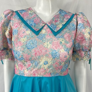 May include: A vintage dress featuring a floral lace bodice in pink, blue, and yellow hues, complemented by a turquoise collar and a matching skirt. The dress has short, puffed sleeves with lace cuffs.