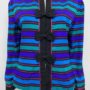 May include: A colorful striped jacket with a round neckline and long sleeves. The jacket features horizontal stripes in shades of blue, teal, and purple, accented with pink lines. Black bows adorn the front, adding a decorative touch. The jacket is a vintage fashion item.