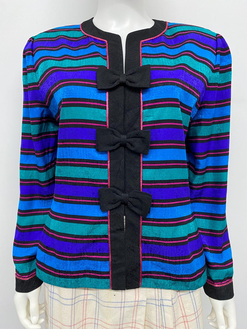 May include: A colorful striped jacket with a round neckline and long sleeves. The jacket features horizontal stripes in shades of blue, teal, and purple, accented with pink lines. Black bows adorn the front, adding a decorative touch. The jacket is a vintage fashion item.