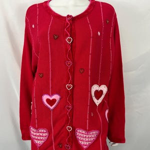 May include: A bright red long-sleeve cardigan featuring a round neckline and a front button closure with heart-shaped buttons. The garment is decorated with various heart-shaped appliqués in shades of pink and red, along with vertical beaded lines.