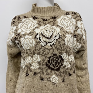 May include: A tan sweater with a mock turtleneck and a floral pattern. The pattern features large white and brown roses with green stems and leaves. The sweater has long sleeves and a ribbed collar.