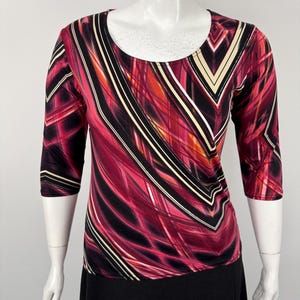 May include: A three-quarter sleeve top with a scoop neckline. The top features a vibrant abstract pattern in shades of red, black, and cream. The bottom portion of the mannequin is wearing a black skirt.