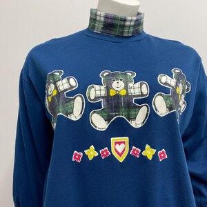 May include: A navy blue sweatshirt with three cartoon teddy bears in plaid patterns. The bears have white outlines and yellow bow ties. The shirt has a plaid turtleneck collar and a row of flowers and a heart graphic.