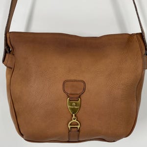 May include: A tan leather shoulder bag featuring a gold-tone metal hardware accent on the front flap. The hardware includes a shield-shaped emblem with the text Etienne Aigner and a horseshoe-shaped clasp. The bag has a matching leather shoulder strap.