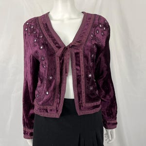 May include: A plum-colored velvet jacket with a V-neck and long sleeves. The jacket features decorative embroidery and small, round, reflective embellishments. A tie closure is at the neckline. The jacket is displayed on a mannequin.