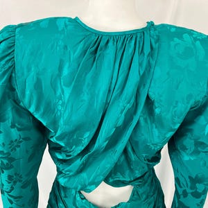 May include: Teal-colored vintage dress with long sleeves and a floral pattern. The dress features a draped back and shoulder pads. The fabric has a subtle sheen, adding to its elegant appearance. A keyhole detail is visible at the back.
