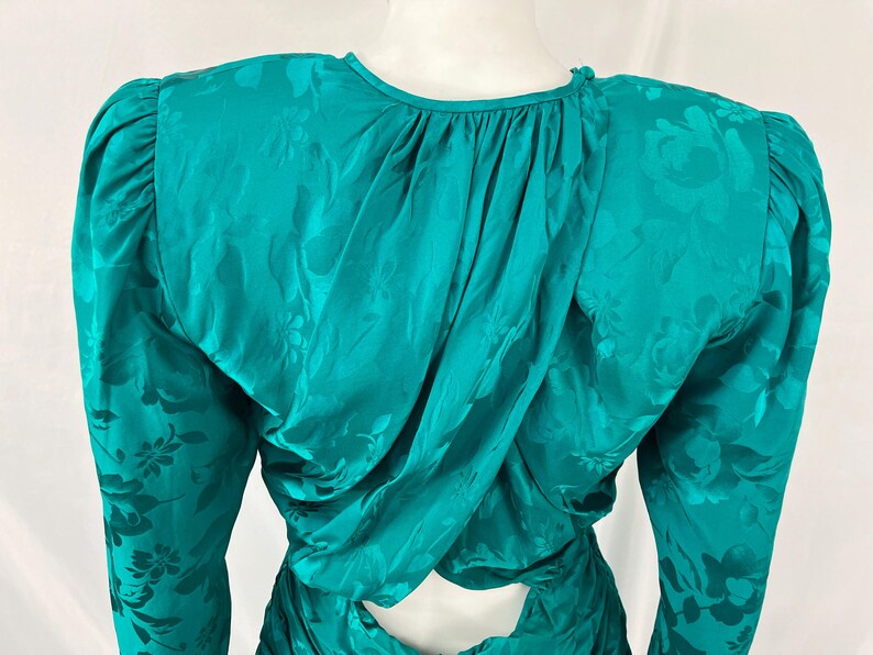 May include: Teal-colored vintage dress with long sleeves and a floral pattern. The dress features a draped back and shoulder pads. The fabric has a subtle sheen, adding to its elegant appearance. A keyhole detail is visible at the back.