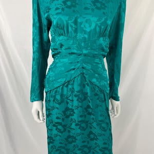 May include: Teal-colored vintage dress with a floral pattern. The long-sleeved dress features a ruched waist and a knee-length skirt. The fabric has a subtle sheen, and the floral design is visible throughout the dress.