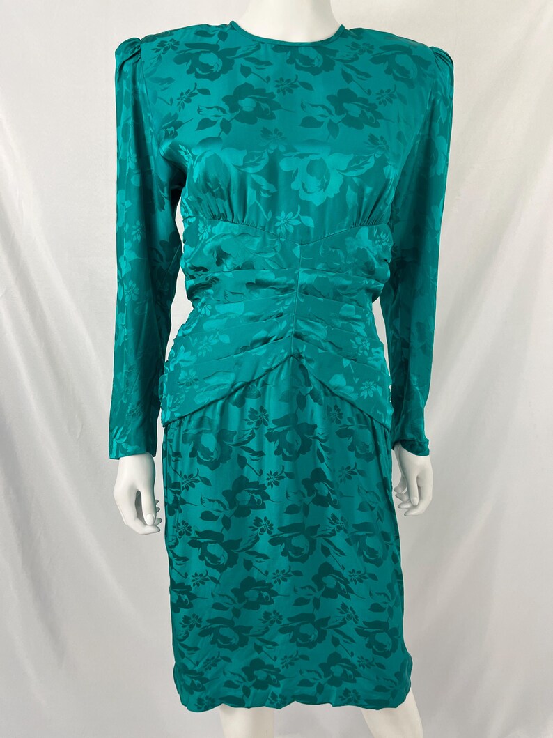 May include: Teal-colored vintage dress with a floral pattern. The long-sleeved dress features a ruched waist and a knee-length skirt. The fabric has a subtle sheen, and the floral design is visible throughout the dress.