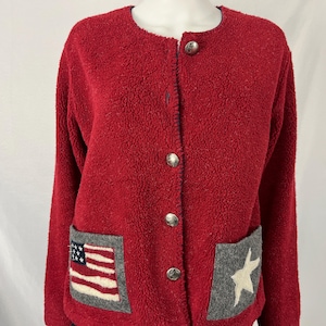 May include: A red fleece jacket with a round collar and silver buttons. The jacket features two front pockets, one with an American flag design and the other with a white star on a gray background. A cozy outerwear garment.