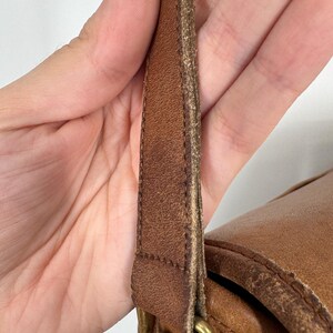 May include: Close-up of a brown leather bag with a shoulder strap. The strap is attached to the bag with a brass-colored metal buckle. The leather shows signs of wear and tear.
