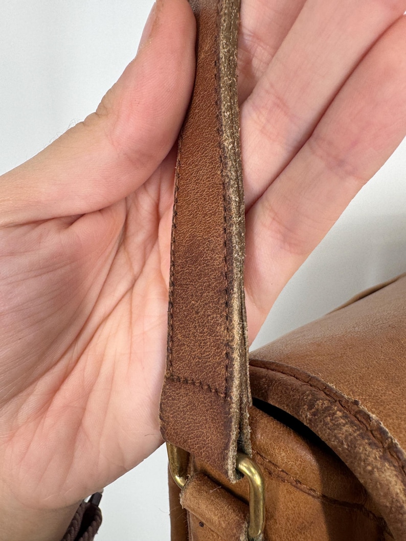 May include: Close-up of a brown leather bag with a shoulder strap. The strap is attached to the bag with a brass-colored metal buckle. The leather shows signs of wear and tear.