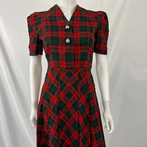 May include: A red and green plaid dress with short sleeves and a v-neck. The dress has a fitted bodice and a full skirt. The dress has two silver buttons on the front.