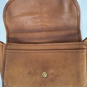 May include: Open view of a vintage brown leather handbag. The bag features a flap closure with a brass clasp and a front pocket. The leather shows signs of wear, adding to its classic appeal. The interior is visible, showcasing the bag's construction.