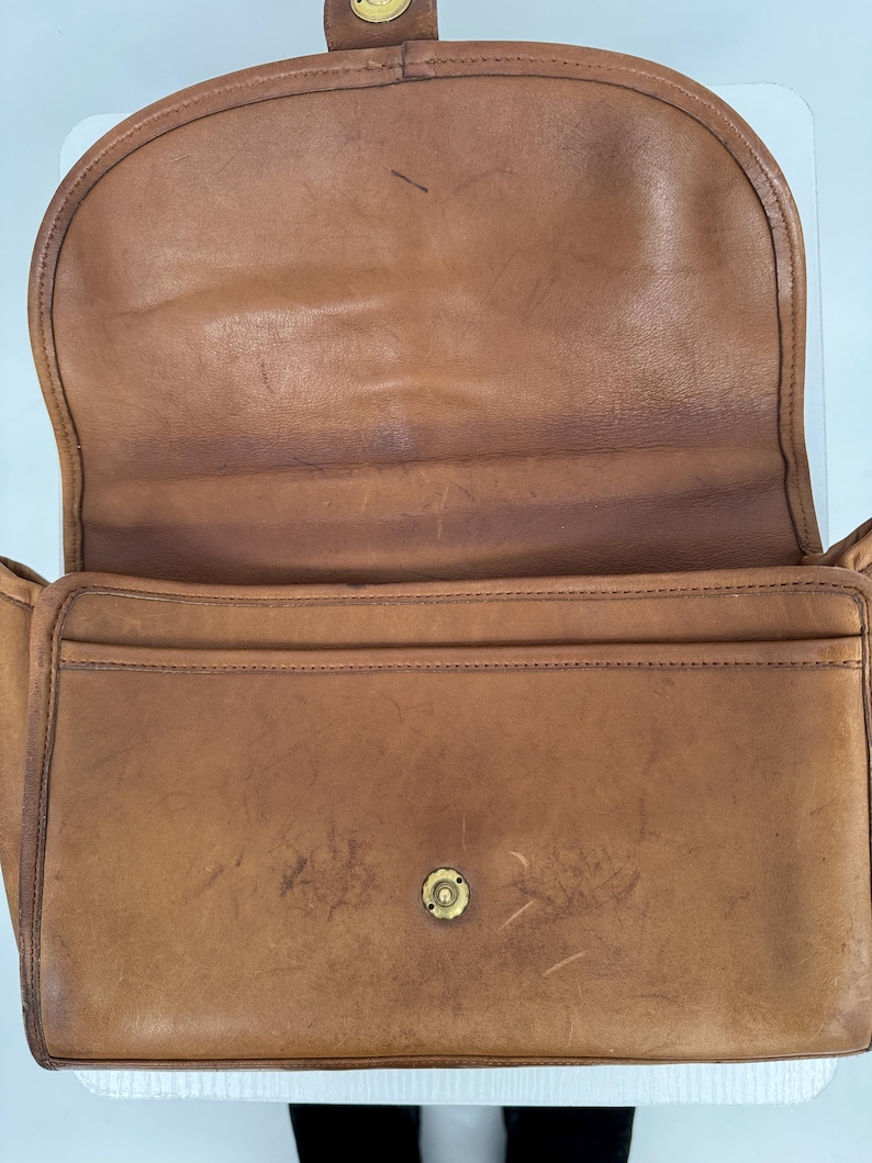 May include: Open view of a vintage brown leather handbag. The bag features a flap closure with a brass clasp and a front pocket. The leather shows signs of wear, adding to its classic appeal. The interior is visible, showcasing the bag's construction.
