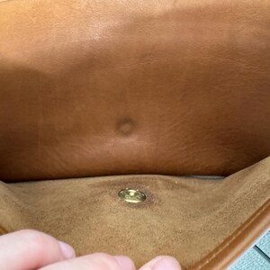 May include: A close-up of the interior of a brown leather bag. The bag has a gold-colored clasp and a small, circular mark on the inside. The leather appears to be worn and has a natural grain.