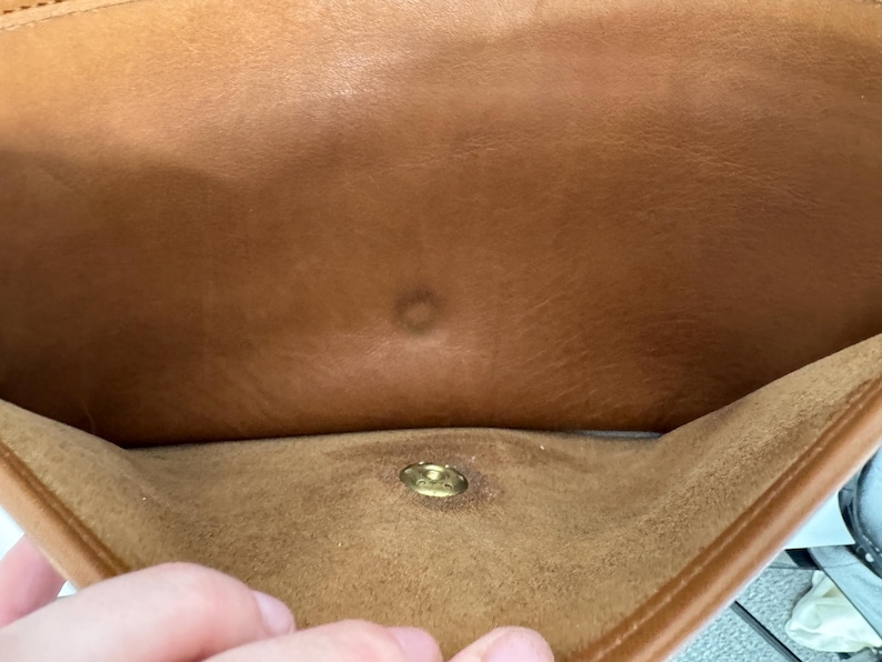 May include: A close-up of the interior of a brown leather bag. The bag has a gold-colored clasp and a small, circular mark on the inside. The leather appears to be worn and has a natural grain.