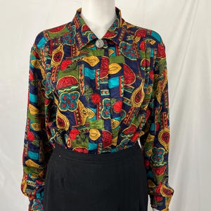 May include: A long-sleeved button-down shirt with a colorful pattern. The shirt features a navy blue background with red, yellow, green, and teal shapes. The collar has a silver button. The shirt is paired with a black skirt.